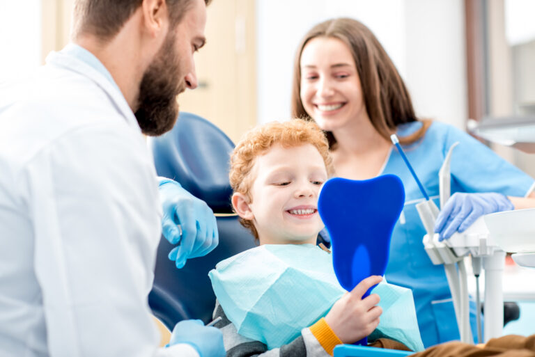 Child smiling in dental chair while holding a tooth-shaped mirror during pediatric dental sealant appointment